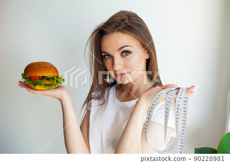 Woman holding unhealthy burger with measuring tape in kitchen. 90228981
