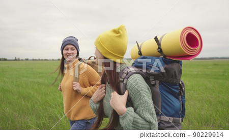 Girl friends with backpacks walk across field hiking 90229914