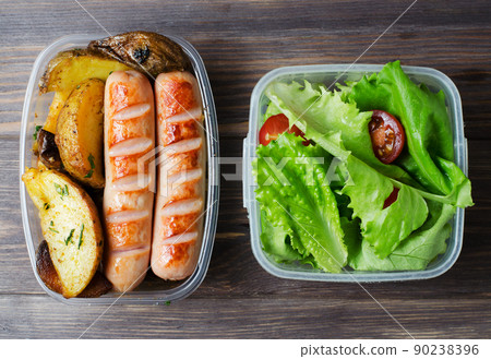 Grilled sausages, potatoes, green lettuce with tomatoes in a plastic box. Unhealthy diet. Wooden background Grilled sausages, potatoes, green lettuce with tomatoes in a plastic box. Unhealthy diet. Wooden background 90238396