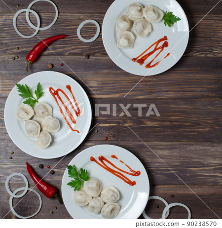 Traditional Russian pelmeni, ravioli, dumplings on a white plate with red sauce and parsley. Dark wooden background. Onion rings, chili. Top view. Copy space Traditional Russian pelmeni, ravioli, dumplings on a white plate with red sauce and parsley. Dark wooden background. Onion rings, chili. Top view. Copy space 90238570