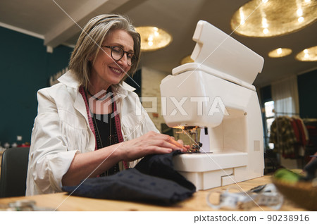 Inspired stylish European mature woman dressmaker sews clothes on sewing machine in a tailoring atelier. Inspired stylish European mature woman dressmaker sews clothes on sewing machine in a tailoring atelier. 90238916