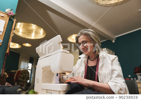 Elderly female fashion designer, dressmaker works, sews clothes on sewing machine. Process of tailoring and repair of clothing in atelier or workshop Elderly female fashion designer, dressmaker works, sews clothes on sewing machine. Process of tailoring and repair of clothing in atelier or workshop 90238919