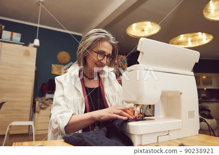 Elegant mature woman clothing designer tailor, sews clothes on sewing machine, stitches fabric, creates new clothes collection in tailoring atelier 90238921