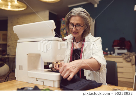 Stylish fashionable clothes designer mature female needlewoman sews at a sewing machine. Manufacturer, tailor in the process of creative development Stylish fashionable clothes designer mature female needlewoman sews at a sewing machine. Manufacturer, tailor in the process of creative development 90238922