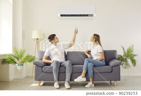 Young couple sitting on couch in living room at home switch on their air conditioner Young couple sitting on couch in living room at home switch on their air conditioner 90239253