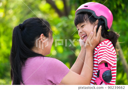 Asian mother helps daughter put on protective pads and safety helmet before practicing roller skating in the park. Exciting outdoor activities for kids. Asian mother helps daughter put on protective pads and safety helmet before practicing roller skating in the park. Exciting outdoor activities for kids. 90239662