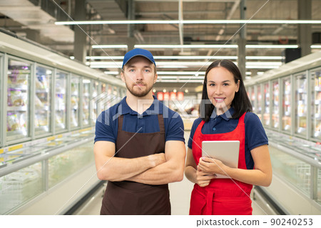 Horizontal medium portrait if man and woman wearing uniform standing together in supermarket aisle looking at camera Horizontal medium portrait if man and woman wearing uniform standing together in supermarket aisle looking at camera 90240253