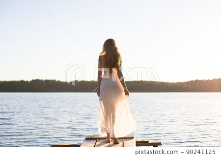 Beautiful Russian girl dressed in a white dress, walking along a wooden pier on the bank of a river or a lake 90241125