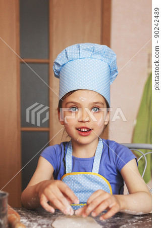 5 year old girl in blue dress and dressed as chef in kitchen prepares pastries from dough 5 year old girl in blue dress and dressed as chef in kitchen prepares pastries from dough 90242489
