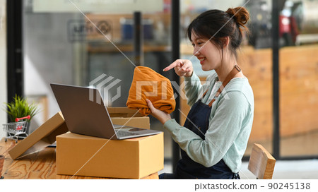 Young female small business owner showing clothes while live streaming online on laptop computer. Online selling, E-commerce 90245138