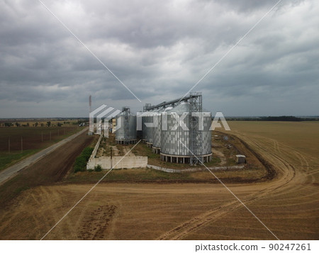 Grain elevator. Metal grain elevator in agricultural zone. Agriculture storage for harvest. Grain elevators on green nature background. Exterior of agricultural factory. Nobody. Grain elevator. Metal grain elevator in agricultural zone. Agriculture storage for harvest. Grain elevators on green nature background. Exterior of agricultural factory. Nobody. 90247261