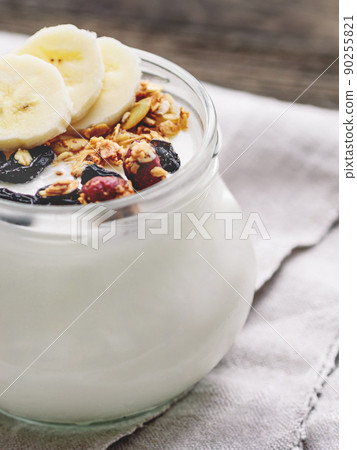 Natural homemade yogurt in a glass jar. Healthy food for breakfast with muesli. Jar with granola and banana slices on linen tablecloth on on wooden table. Natural homemade yogurt in a glass jar. Healthy food for breakfast with muesli. Jar with granola and banana slices on linen tablecloth on on wooden table. 90255821