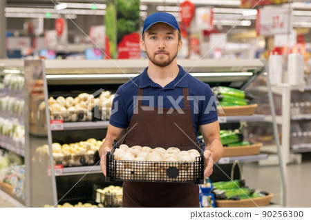 Vertical medium long shot of modern store worker wearing uniform holding box full of fresh mushrooms 90256300