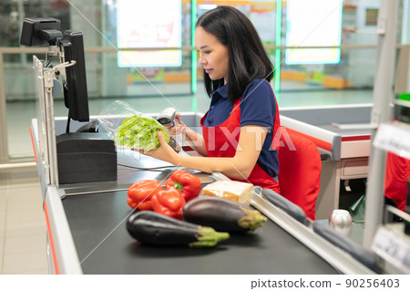 attractive Asian cashier wearing red apron selling fresh vegetables to customer 90256403