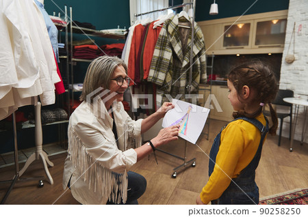 Pleasant mature European woman, cheerful caring and loving grandmother admiring the drawing - clothes sketch- of her granddaughter adorable baby girl while working in a fashion design studio Pleasant mature European woman, cheerful caring and loving grandmother admiring the drawing - clothes sketch- of her granddaughter adorable baby girl while working in a fashion design studio 90258250
