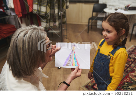 Loving grandma, European elderly woman smiles to her charming granddaughter and admires her beautiful sketch of clothes. Fashion design concept, art, creativity, hobby Loving grandma, European elderly woman smiles to her charming granddaughter and admires her beautiful sketch of clothes. Fashion design concept, art, creativity, hobby 90258251