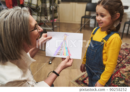 Pleasant elderly Caucasian woman tailor, caring and loving granny smiles admiring the clothes sketch of her adorable granddaughter while working in a fashion design studio and tailoring atelier Pleasant elderly Caucasian woman tailor, caring and loving granny smiles admiring the clothes sketch of her adorable granddaughter while working in a fashion design studio and tailoring atelier 90258252