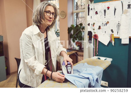 Pleasant charming mature woman seamstress tailor ironing the blue shirt from her new collection in a sewing workshop. Sewing process, fashion design, creation of new garment in tailoring atelier Pleasant charming mature woman seamstress tailor ironing the blue shirt from her new collection in a sewing workshop. Sewing process, fashion design, creation of new garment in tailoring atelier 90258259