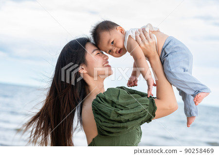 Happy Joyful mother and Son having fun Playing on the Beach together. Asian mom walks by ocean on the beach and throwing up her son in the air at the tropical view. Motherhood Family Concept. 90258469