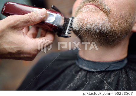 Barber master is shaving of handsome mature bearded man in salon. Hair artist making hairstyle for person in barbershop. Services of professional stylist. Fashion haircare for men 90258567