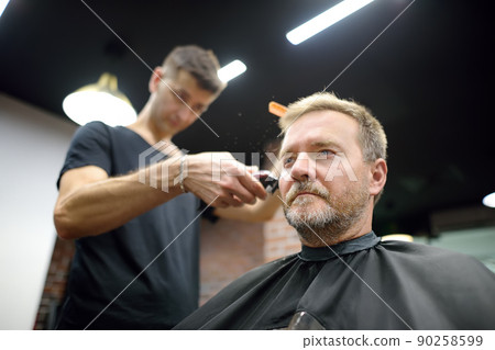 Hairdresser is cutting hair of handsome bearded mature man in salon. Stylist making hairstyle with electric shaver for person in barbershop. Services of a professional stylist. Fashion haircut 90258599