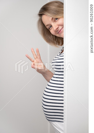 Pregnant woman peeking out of white wall and showing three fingers sign. Happy Pregnant Woman looking at camera and smiling. Baby expectation. Pregnant woman peeking out of white wall and showing three fingers sign. Happy Pregnant Woman looking at camera and smiling. Baby expectation. 90261009