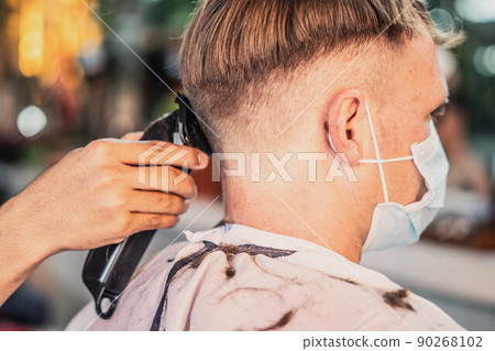 Lifestyle photo barbershop. Young man customer wears face mask, side back rear view. Hairdresser male hand work move comb tool electric clipper. Modern short hair locks cut. Fashion service salon Lifestyle photo barbershop. Young man customer wears face mask, side back rear view. Hairdresser male hand work move comb tool electric clipper. Modern short hair locks cut. Fashion service salon 90268102