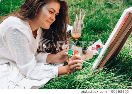 Artist painting on the easel outdoors in the garden. Open air outdoor art workshop. Draw on the canvas with brush and palette sitting on the grass during a picnic in a park Artist painting on the easel outdoors in the garden. Open air outdoor art workshop. Draw on the canvas with brush and palette sitting on the grass during a picnic in a park 90268395
