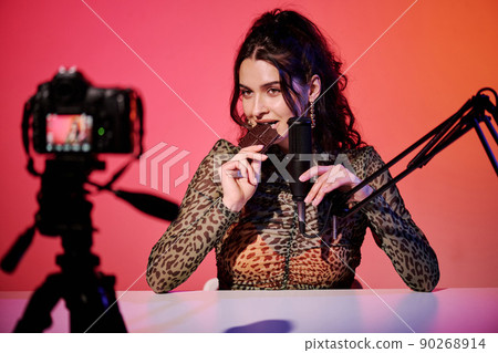 Attractive young Caucasian woman sitting in studio in pink and orange neon light recording chocolate bar eating sounds and video for ASMR vlog 90268914