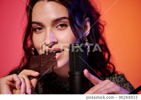 Horizontal close-up of young Caucasian woman sitting in studio in pink and orange neon light recording chocolate bar eating sounds for ASMR vlog 90268915