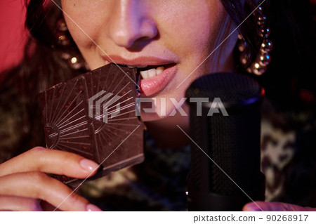 Close-up of young female blogger sitting in studio recording chocolate bar biting and eating sounds for ASMR vlog Close-up of young female blogger sitting in studio recording chocolate bar biting and eating sounds for ASMR vlog 90268917