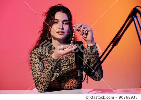 Modern young female blogger sitting in studio in pink and yellow neon light recording sounds for ASMR vlog using bead necklace Modern young female blogger sitting in studio in pink and yellow neon light recording sounds for ASMR vlog using bead necklace 90268933