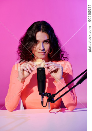 Attractive young female blogger sitting at desk in studio in pink neon light making sounds with crunchy crisps for ASMR vlog, vertical shot 90268955