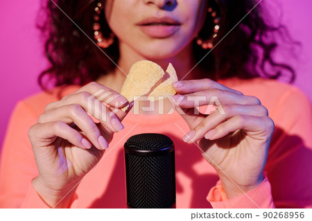 Young female blogger sitting in studio in pink neon light making sounds with crunchy crisps for ASMR vlog 90268956