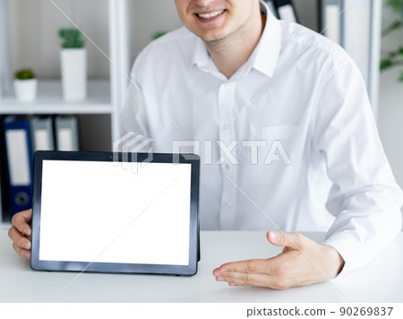 Online presentation. Office man. Computer mockup. Unrecognizable smiling guy pointing to tablet computer with blank screen sitting desk in light room interior. Online presentation. Office man. Computer mockup. Unrecognizable smiling guy pointing to tablet computer with blank screen sitting desk in light room interior. 90269837