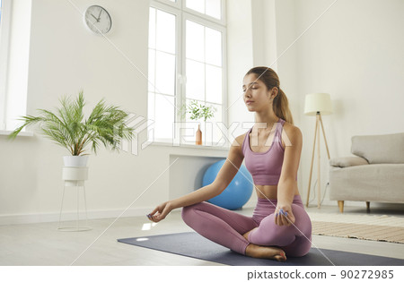 Relaxed and calm young woman at home practicing lotus asana sitting in Suhasana pose. 90272985