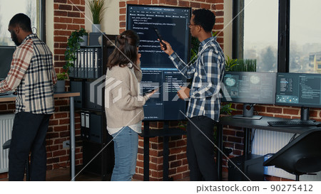 Team of database admins analyzing source code on wall screen tv comparing errors using digital tablet in busy server room. Two cloud programers debugging algorithm in software innovation office. 90275412