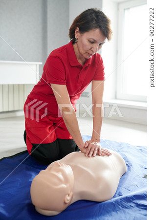 Mature female medical worker in uniform using dummy learning to do heart massage on floor 90277222