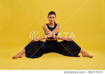 Sporty girl sitting with stretching legs, looking at camera indoors. Slender woman with ponytail holding crossed arms on wide apart knees, isolated on yellow studio background. Concept of exercising. Sporty girl sitting with stretching legs, looking at camera indoors. Slender woman with ponytail holding crossed arms on wide apart knees, isolated on yellow studio background. Concept of exercising. 90277619