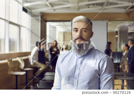 Portrait of serious business man standing in office hall against against background of colleagues. 90278126