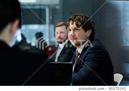 Young businessman in dark suit listening to speaker while sitting at table with his colleagues at meeting Young businessman in dark suit listening to speaker while sitting at table with his colleagues at meeting 90278141