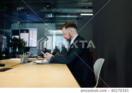 Young businessman in formal suit sitting at table in boardroom and using mobile phone awaiting meeting 90278165