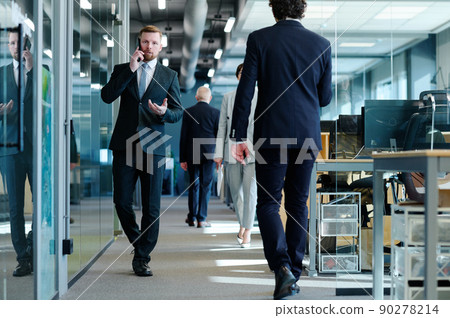 Young businessman in suit talking on mobile phone while walking along office corridor among business people Young businessman in suit talking on mobile phone while walking along office corridor among business people 90278214