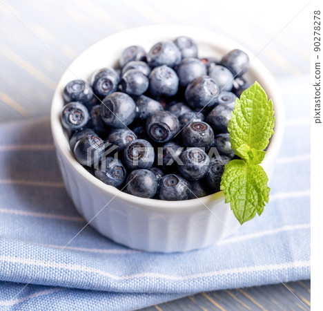 Forest blueberries with fresh mint leaf in white bowl on blue striped napkin. Blueberry - natural superfood. Healthy food. Natural vitamin for eyes. Square photo. Close up. 90278278
