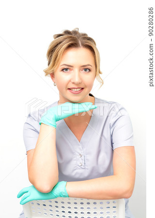 Close-up portrait of young smiling female caucasian healthcare worker sitting and staring at the camera wearing gloves on white background Close-up portrait of young smiling female caucasian healthcare worker sitting and staring at the camera wearing gloves on white background 90280286