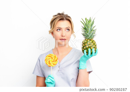 Young caucasian female doctor nutritionist holding fresh pineapple with lollipop over white background Young caucasian female doctor nutritionist holding fresh pineapple with lollipop over white background 90280287
