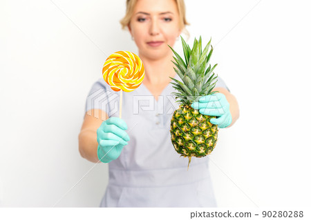 Young caucasian female doctor nutritionist holding fresh pineapple with lollipop over white background Young caucasian female doctor nutritionist holding fresh pineapple with lollipop over white background 90280288