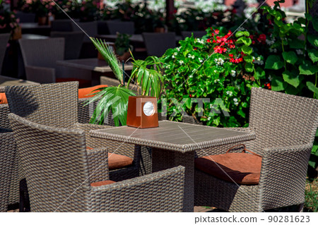 wicker rattan plastic fittings on terrace of street restaurant lit by sun, square table with napkin holder and chairs with soft cushion nearby green plants flowers and palm leaves, close-up, nobody. wicker rattan plastic fittings on terrace of street restaurant lit by sun, square table with napkin holder and chairs with soft cushion nearby green plants flowers and palm leaves, close-up, nobody. 90281623