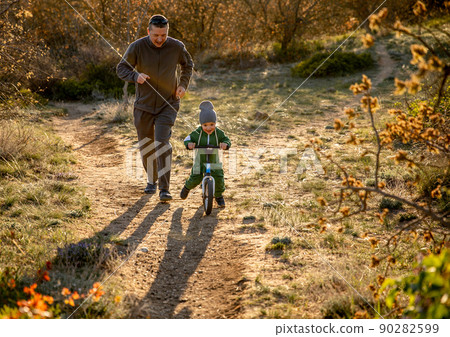 Father and his little son on a balance bike, walking fun in the park. Father and his little son on a balance bike, walking fun in the park. 90282599
