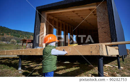 Boy toddler playing as builder on construction site. Back view of child carpenter learning to build wooden frame house in Scandinavian style barnhouse outdoor on sunny day. Carpentry, workshop concept Boy toddler playing as builder on construction site. Back view of child carpenter learning to build wooden frame house in Scandinavian style barnhouse outdoor on sunny day. Carpentry, workshop concept 90282753